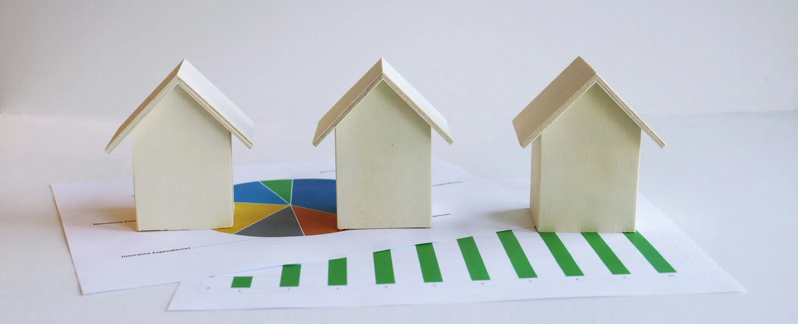 Three small wooden house models on financial charts with green bar and colorful pie graphs on white background
