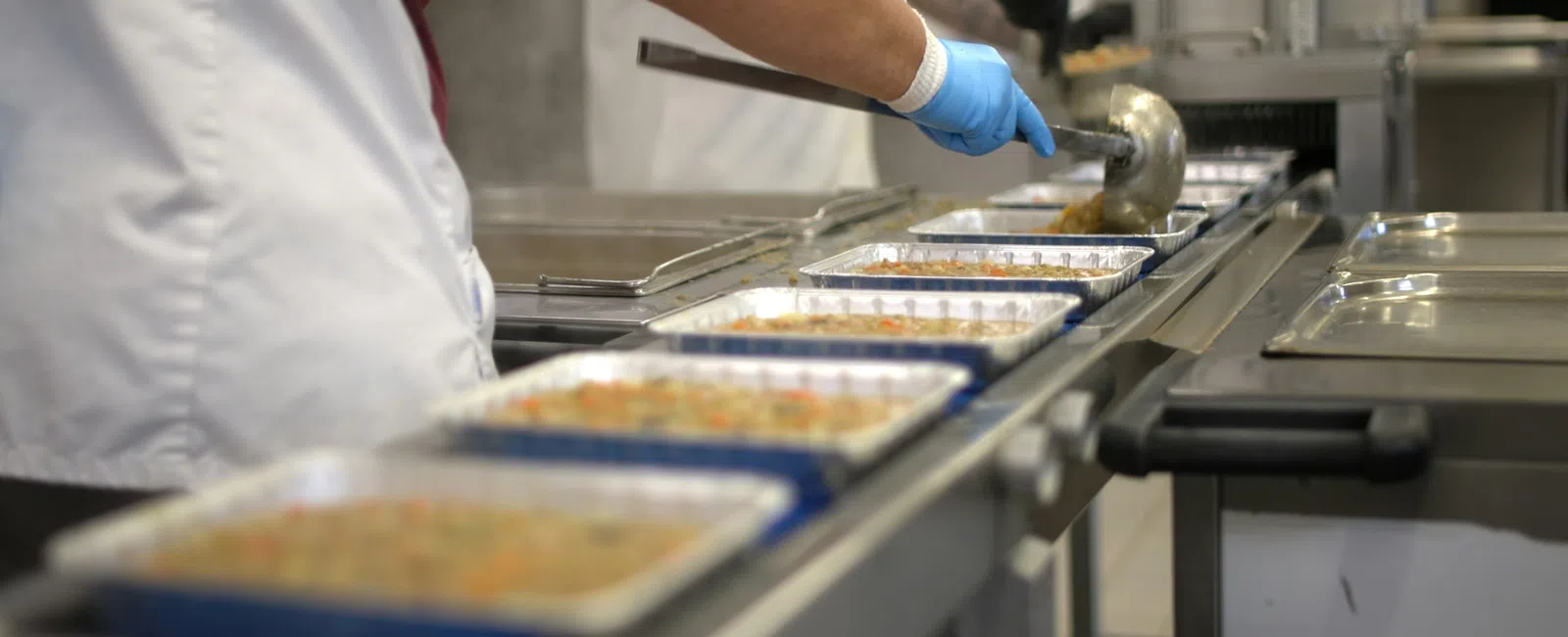 Catering worker serves food into aluminum trays on a production line in a commercial kitchen setting.