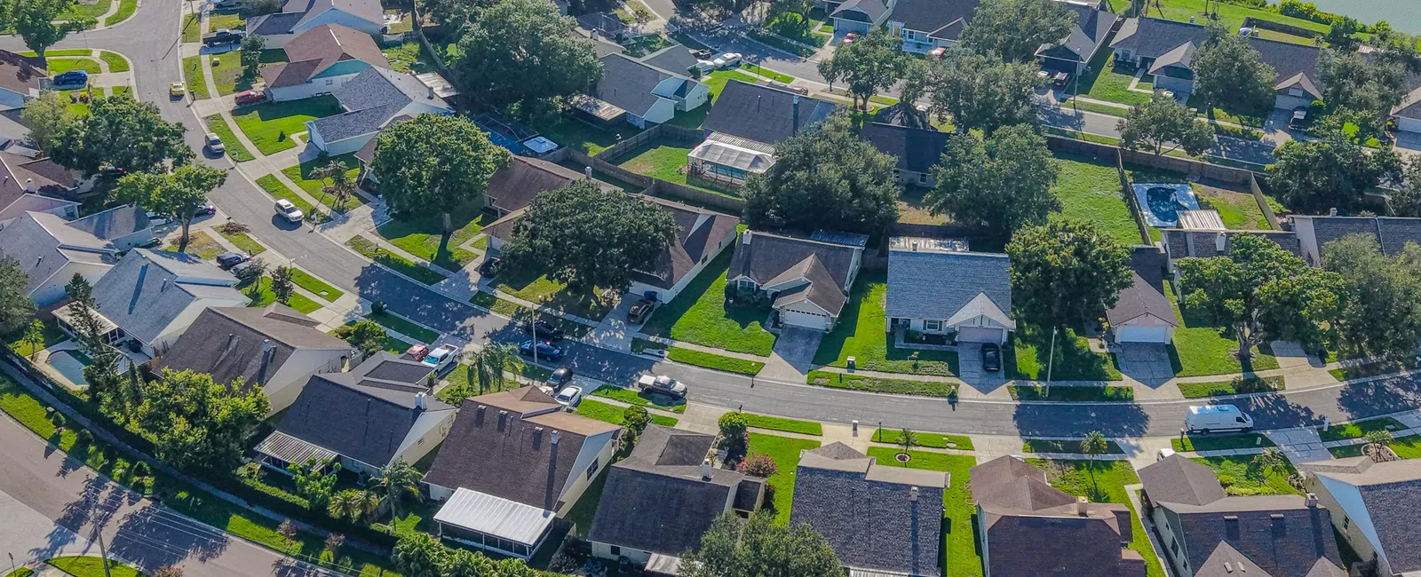 Aerial view of a suburban neighborhood with houses, green lawns, trees, and a small pond in the background.