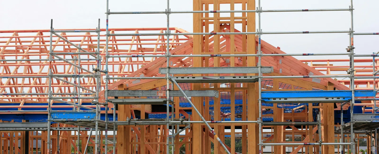 Wooden house frame under construction surrounded by metal scaffolding against a clear sky.