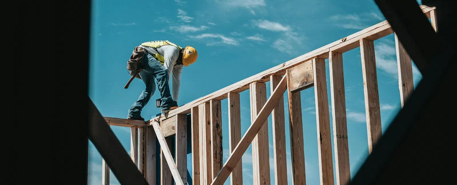 Construction worker in safety gear working on wooden frame structure under clear blue sky