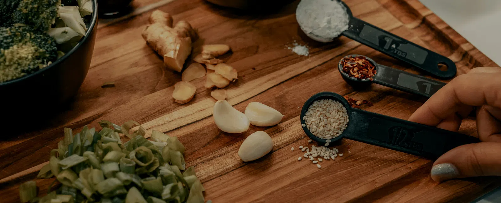 Fresh broccoli, ginger, garlic, chopped scallions, and measuring spoons with sesame seeds and spices on a wooden board.