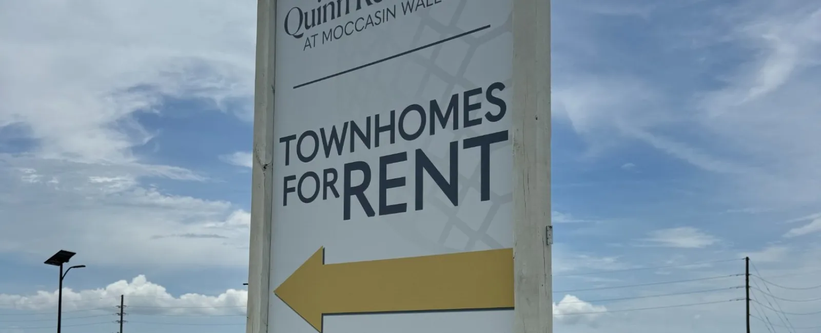 Quinn Residences townhomes for rent sign with arrow and website Live-Moccasin.com on grass roadside.