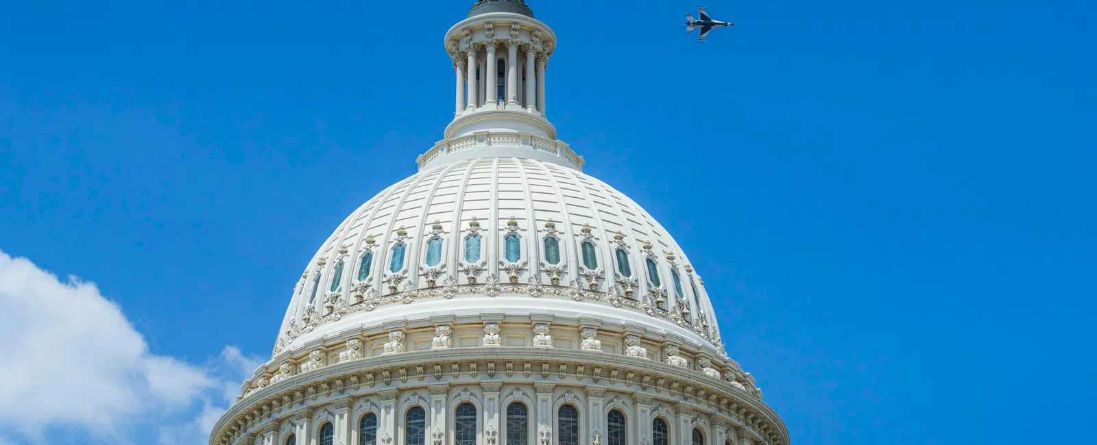 The United States Capitol dome with a military jet flying in clear blue sky above the American flag.