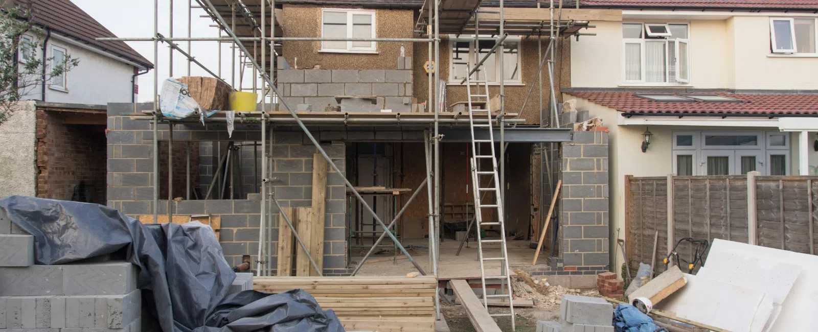 House under construction with scaffolding, bricks, wooden planks, and construction materials in backyard.