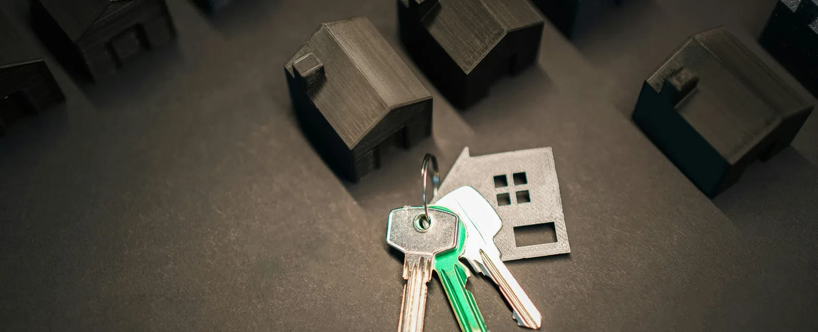 House keys with a house-shaped keychain lying on a dark surface surrounded by small model houses.