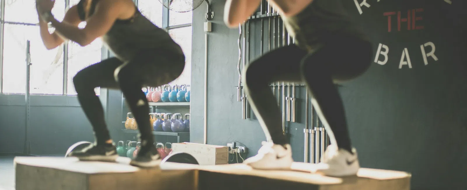 Two women performing box jumps in a gym with kettlebells and motivational wall text behind them.