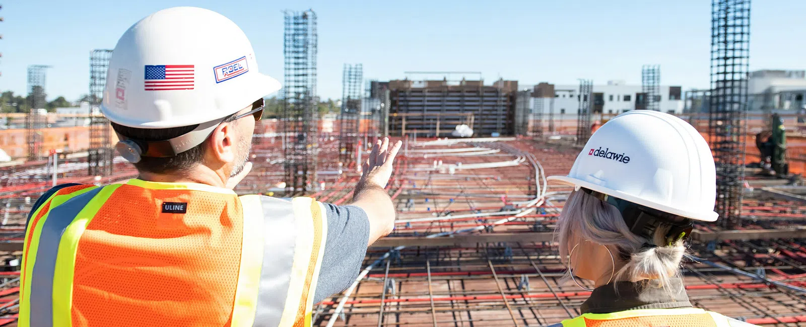 Two construction workers in safety vests and helmets inspecting a large building foundation on a sunny day.