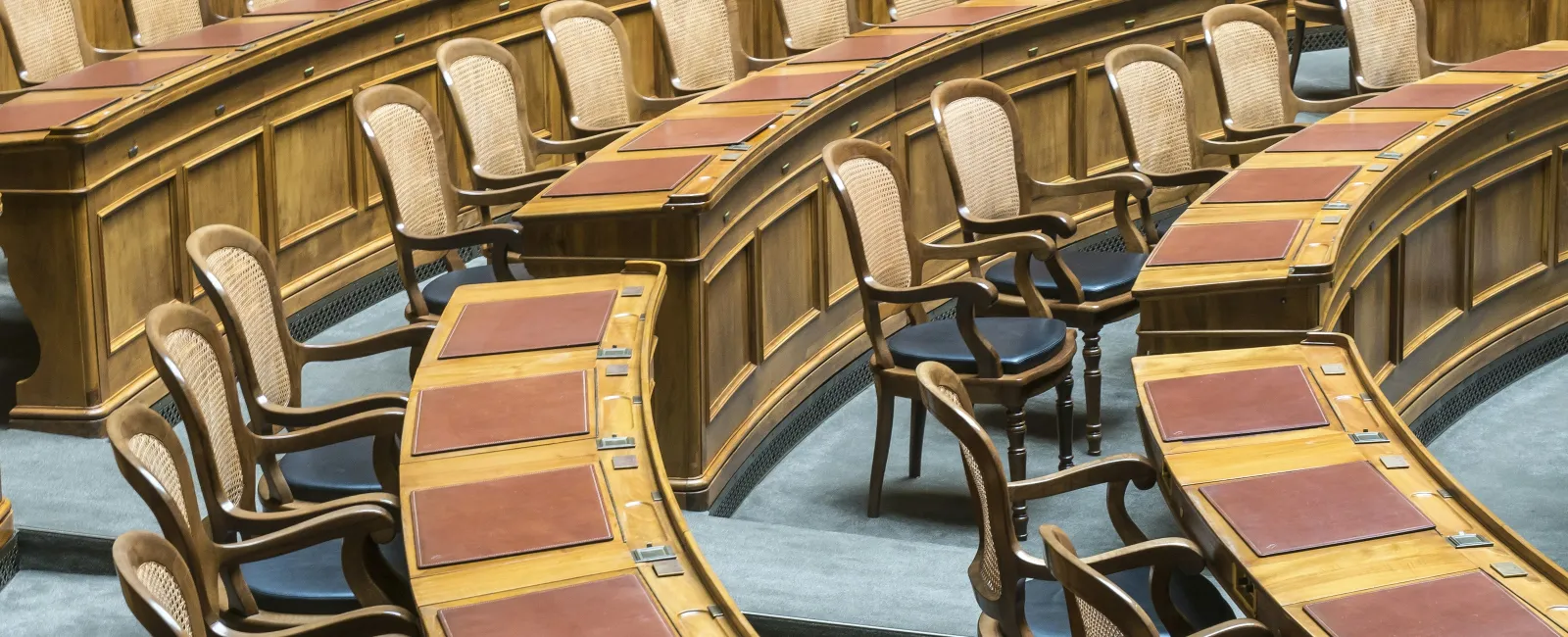 Curved wooden desks with leather pads and matching chairs arranged in a legislative chamber seating area.