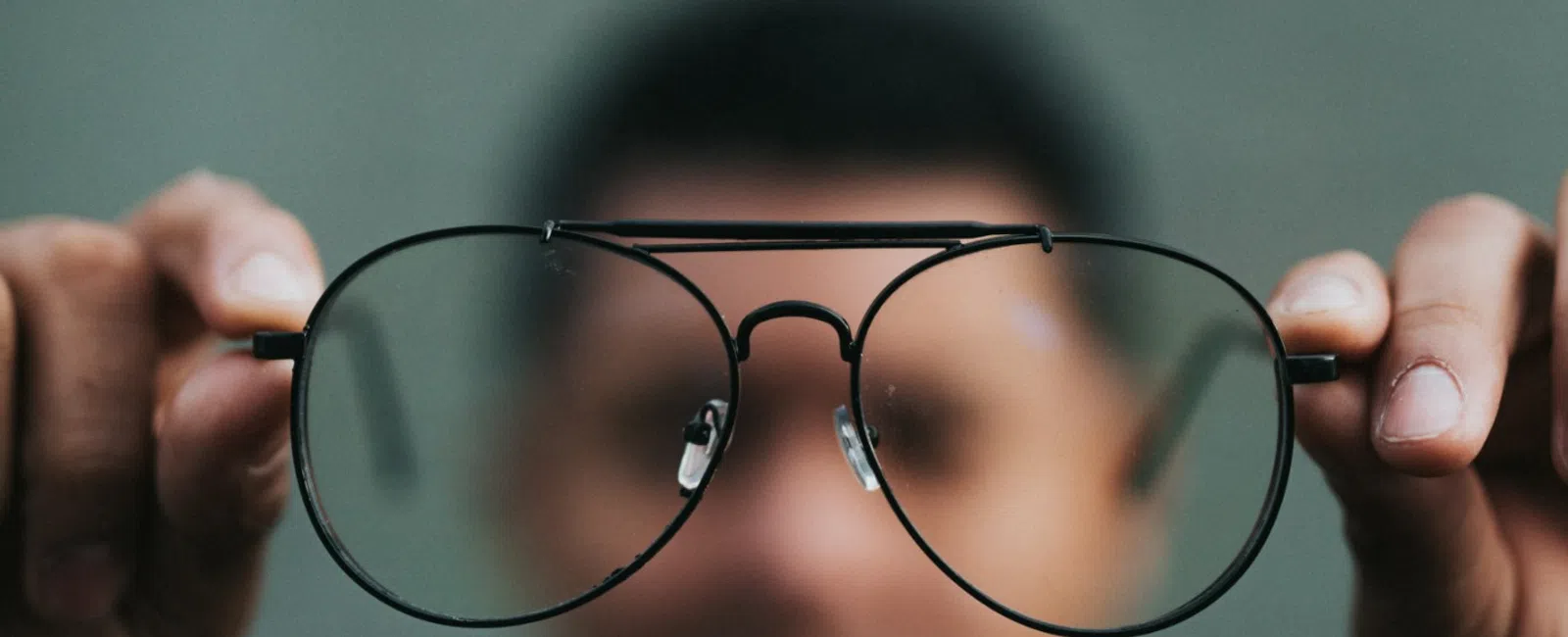 Man holding black-rimmed eyeglasses close to the camera with blurred face in background