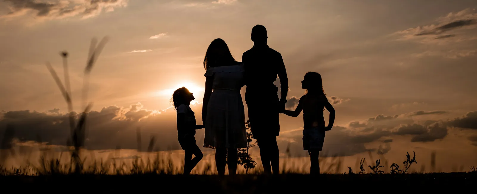 Silhouetted family of four standing together during a sunset with a dramatic cloud-filled sky.