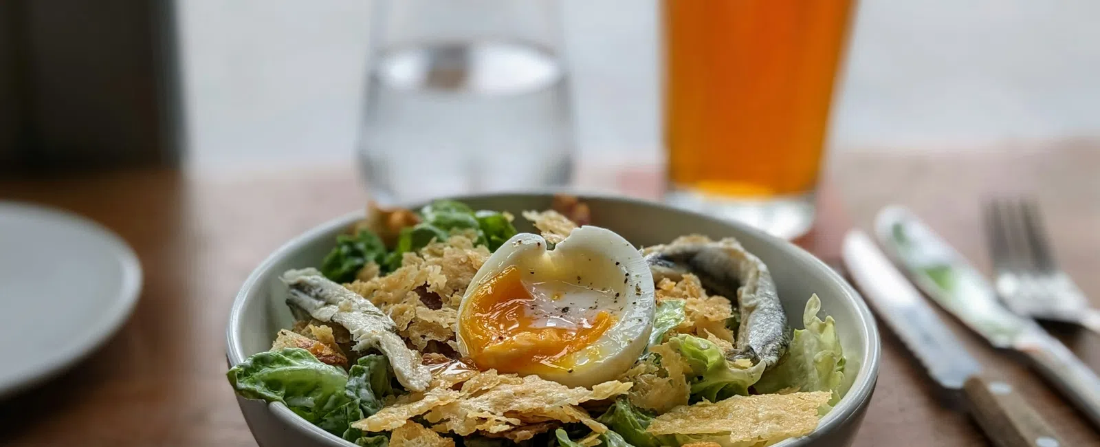 Fresh salad bowl with leafy greens, crispy toppings, and a soft-boiled egg, served with a glass of beer and water.