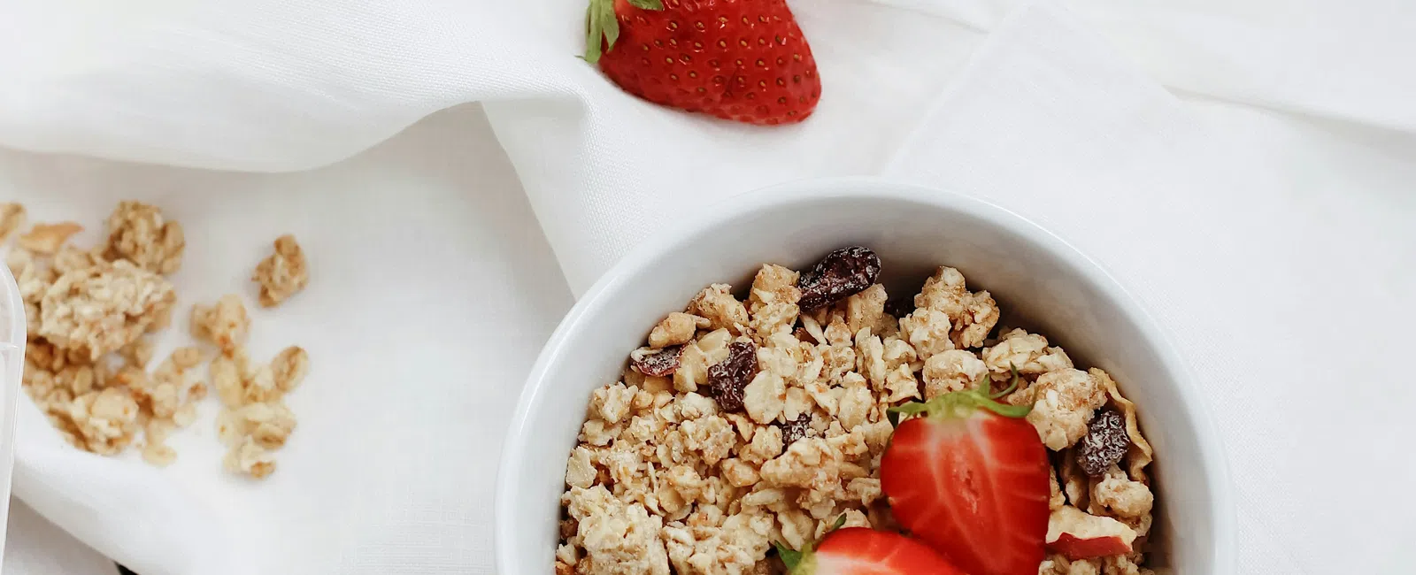 Bowl of granola topped with fresh strawberries on a wooden board with more strawberries and white cloth