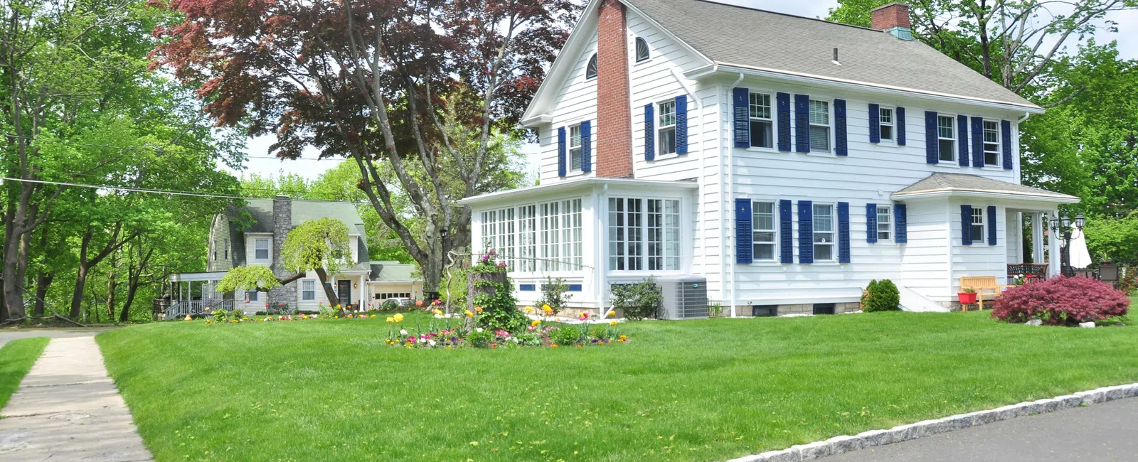 White two-story house with blue shutters surrounded by green lawn, trees, and a colorful flower bed under a blue sky.