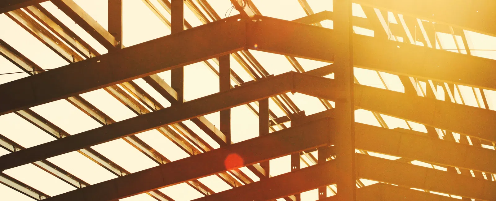 Steel framework of a building under construction with sunlight shining through the beams at sunset.
