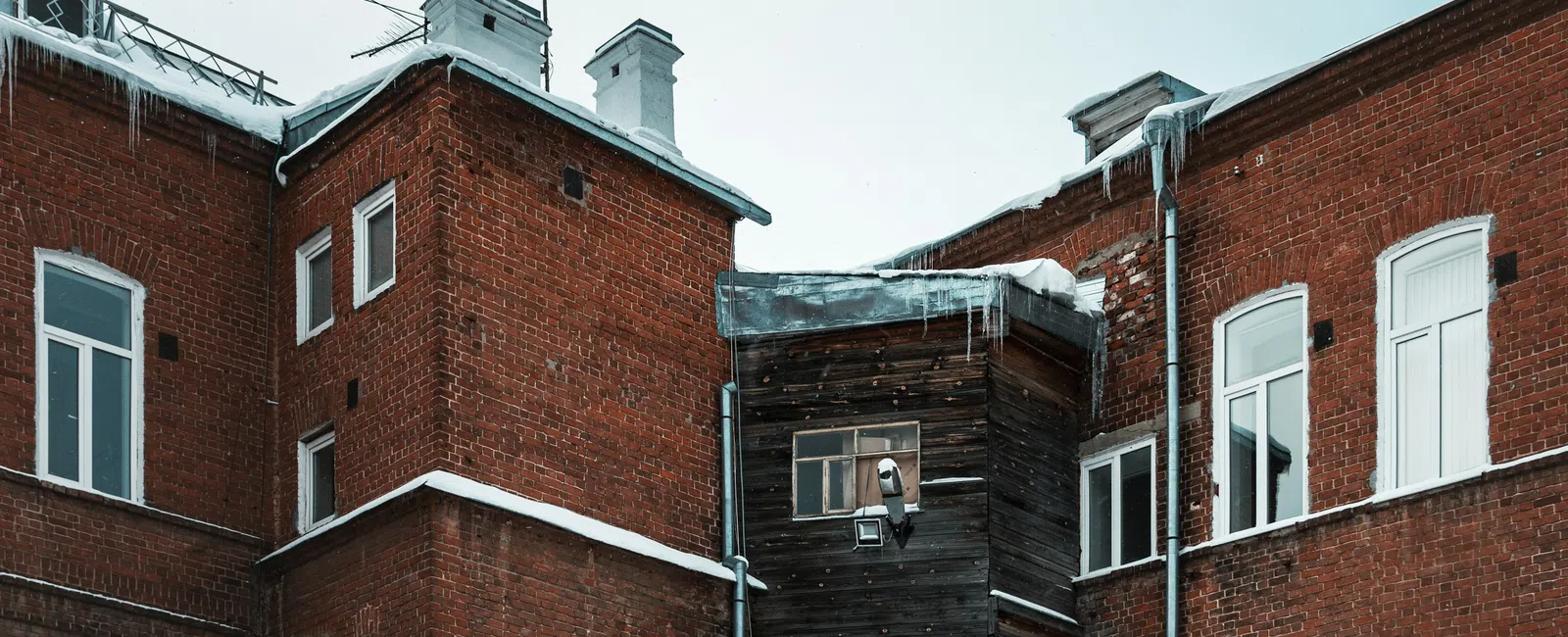 Snow-covered red brick building with wooden bay window extension and icy roofscape under a pale winter sky.
