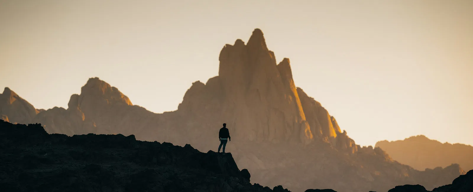Silhouette of person standing on rocky hill with sharp mountain peaks lit by golden hour sunlight