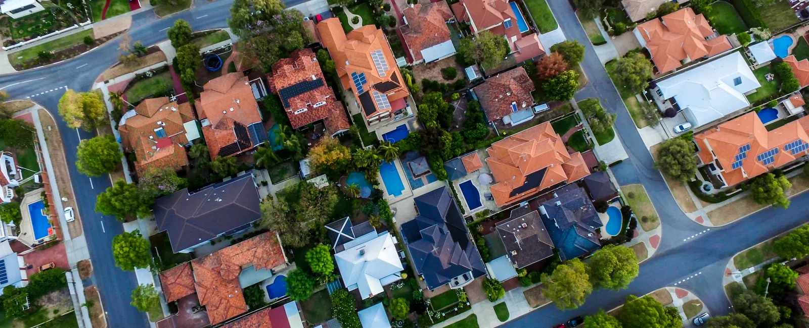 Aerial view of a suburban neighborhood with houses, roads, trees, and swimming pools on a clear day.