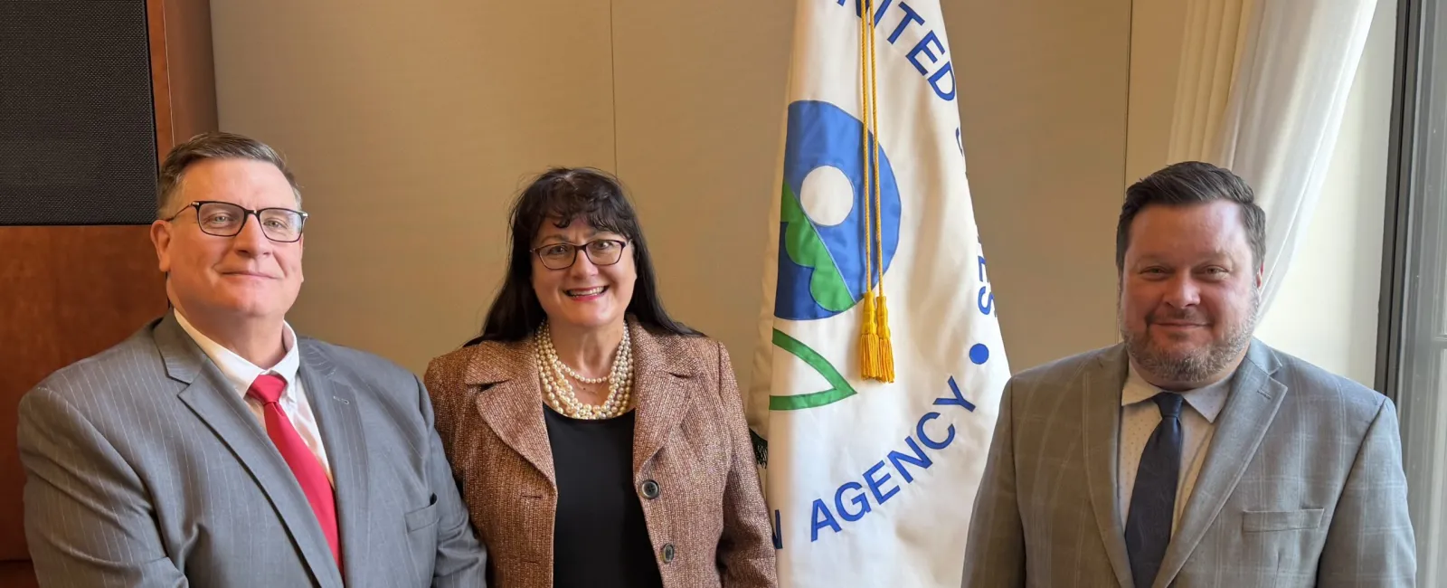 Three professionals posing indoors near a United States EPA flag, dressed in business attire, smiling at camera.
