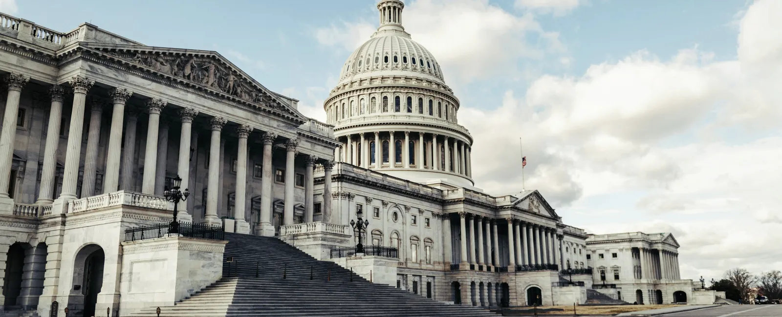 Wide view of the US Capitol building with its dome, stairs, columns, and cloudy blue sky in the background
