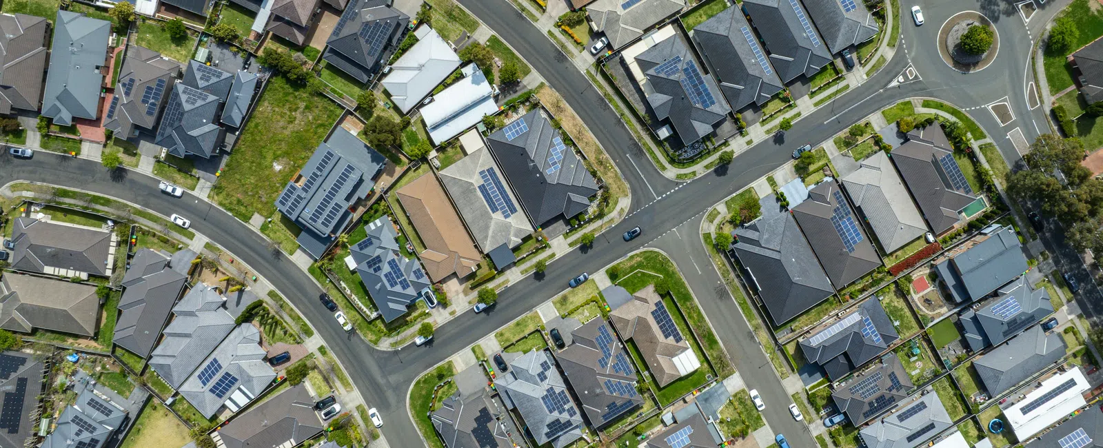 Aerial view of suburban neighborhood with houses, solar panels on roofs, roads, and green yards on a sunny day.