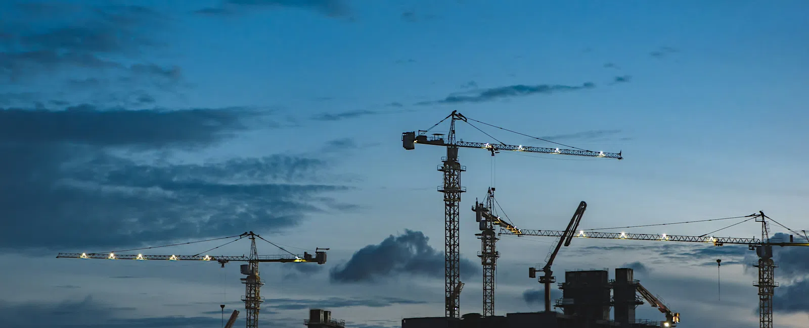Construction cranes illuminated at dusk over a building site with a mountainous backdrop under a blue sky.