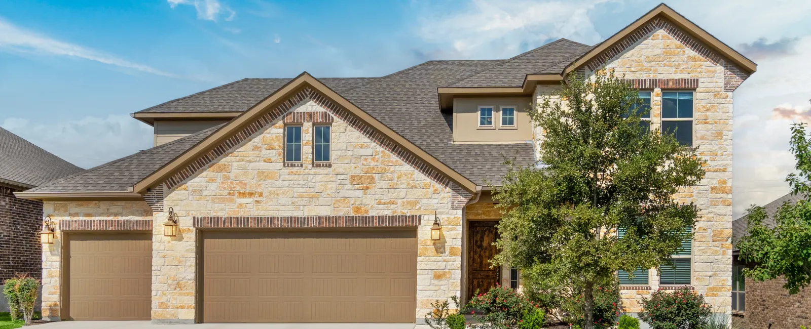Modern two-story house with stone facade, triple garage doors, manicured lawn, and clear blue sky.