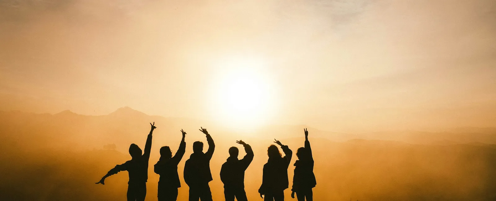 Silhouettes of six people raising hands at sunset on a hill with a golden sky and distant mountains.