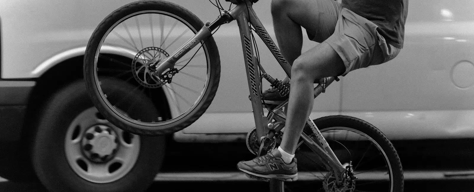 Person performing a wheelie on a bicycle in front of a parked white van on the street