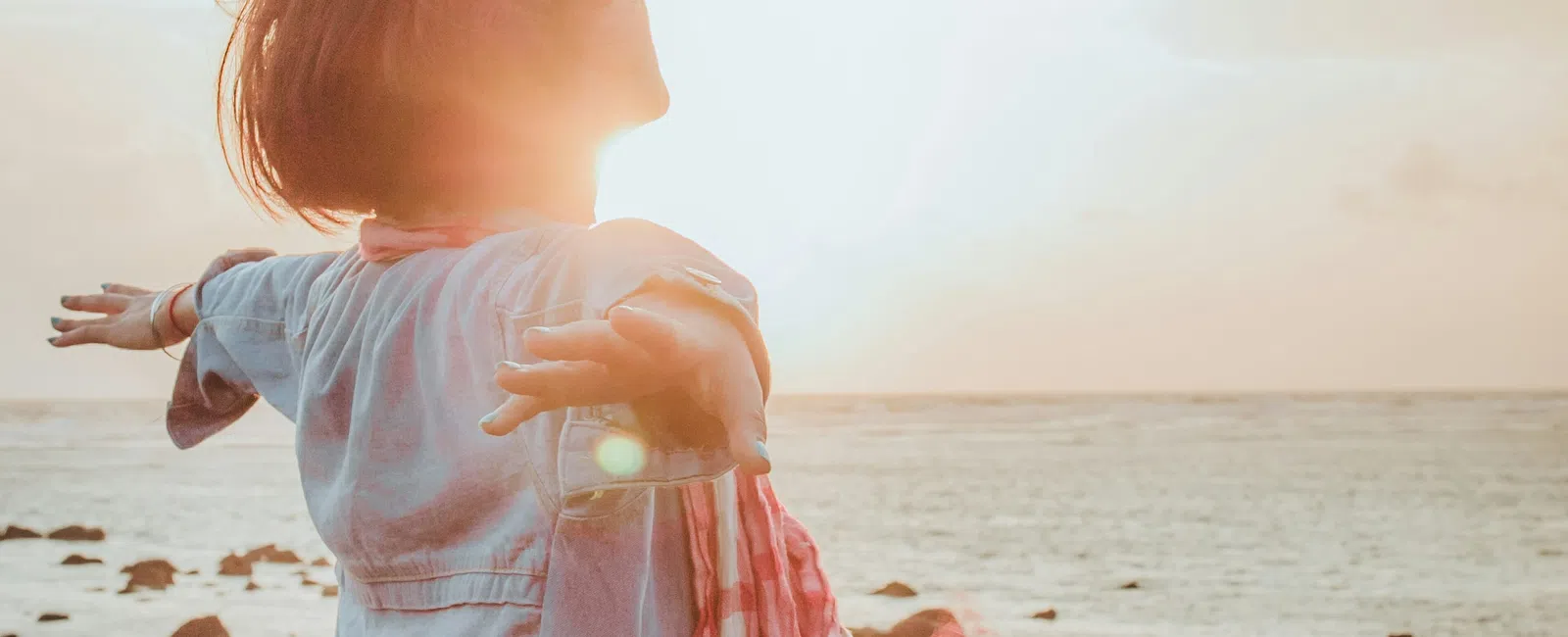 Person with arms outstretched enjoying sunset by rocky sea shore with scarf flowing in breeze.