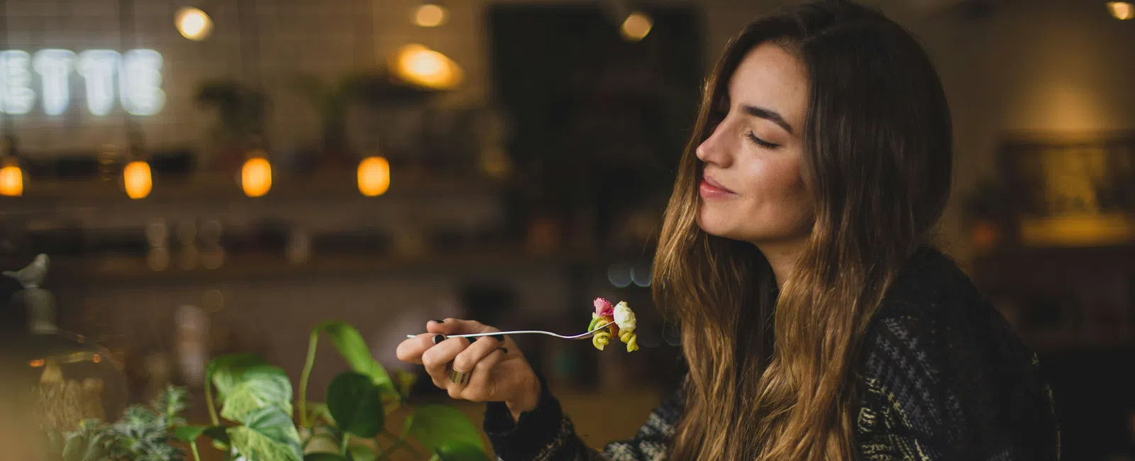 Woman enjoying a meal at a cozy cafe with plants and warm ambient lighting.