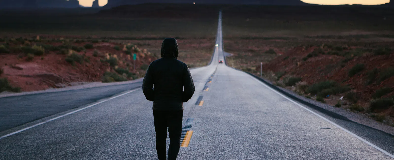 Person walking alone on a long empty road toward desert mesas at dusk with clear sky.