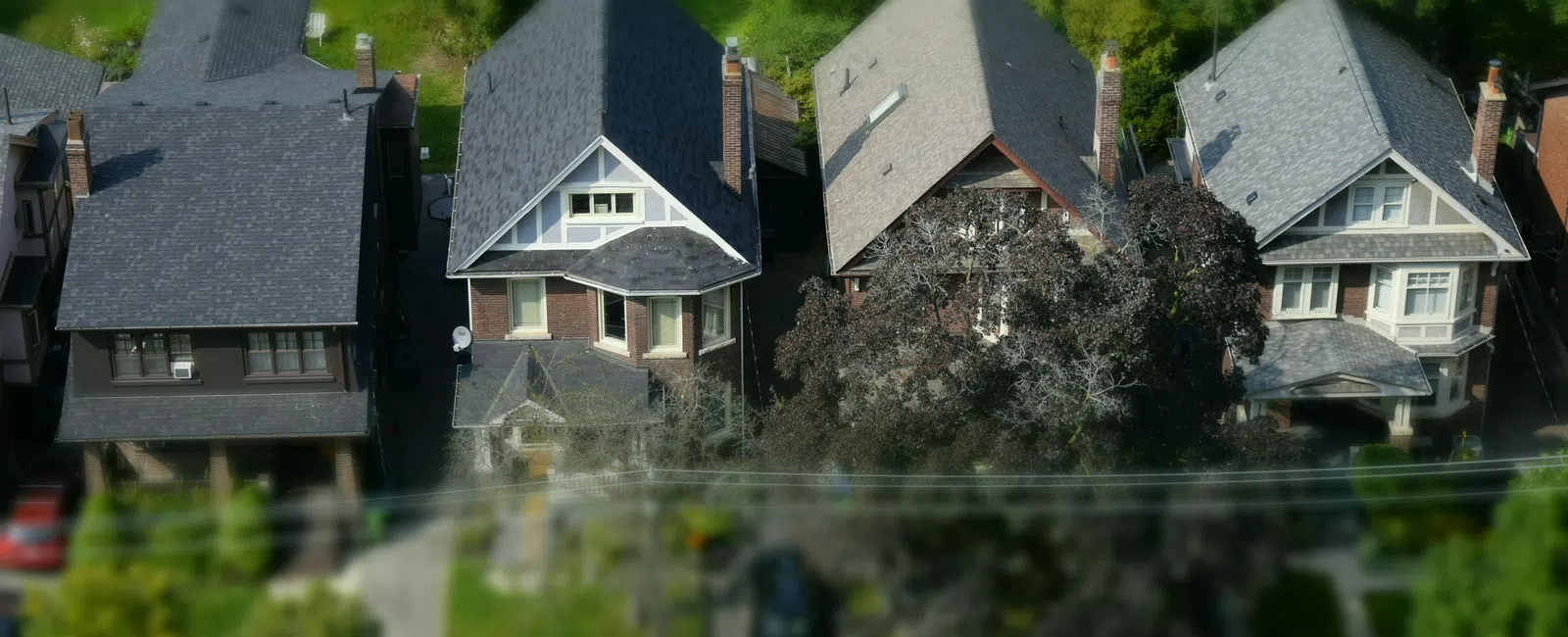 Aerial view of four suburban houses with pitched roofs surrounded by green trees and lawns on a sunny day.