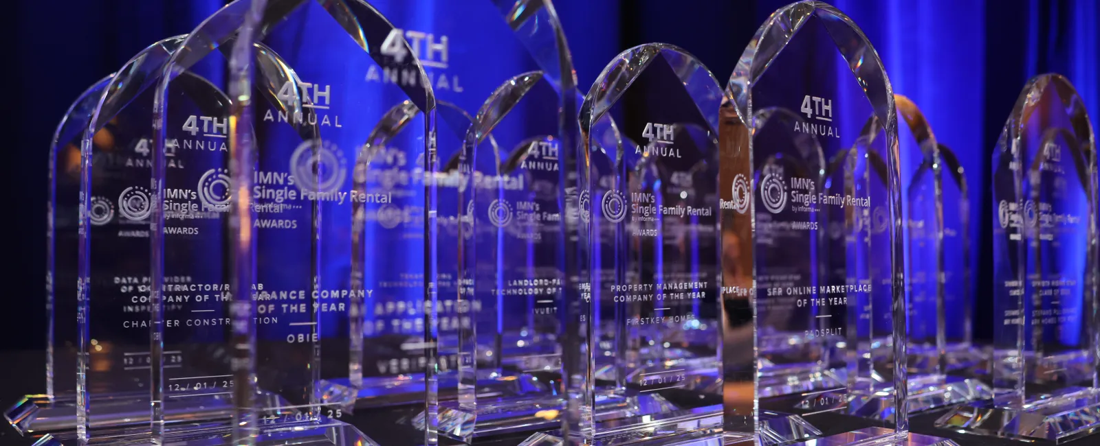 Row of crystal awards for IMN's 4th Annual Single Family Rental event under blue lighting on a black table.