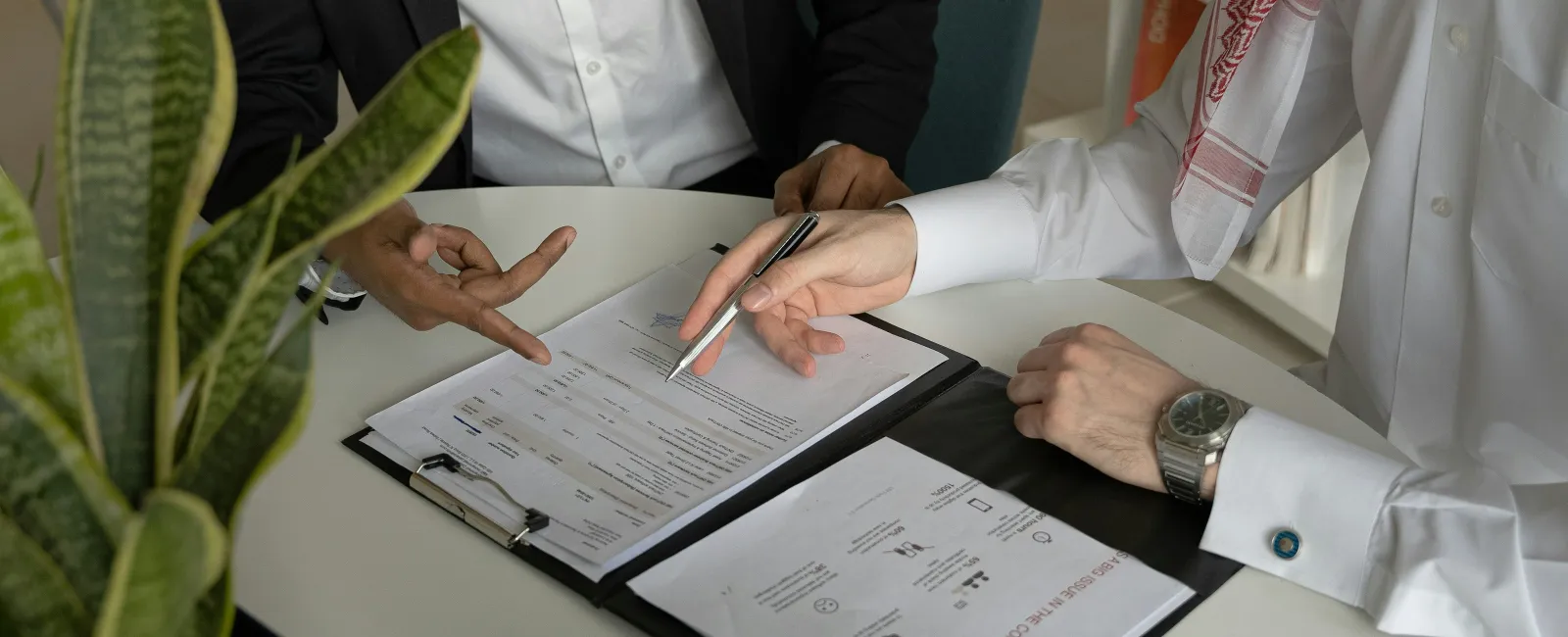 Two men in business attire reviewing documents on a clipboard during a meeting at a white table.
