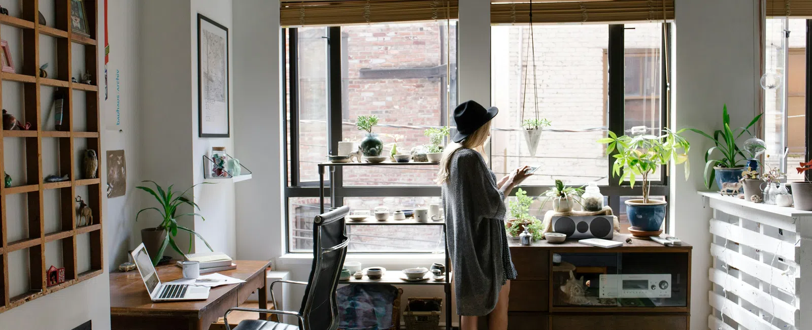 Woman in hat standing by large windows in modern cozy room filled with plants and wooden furniture.