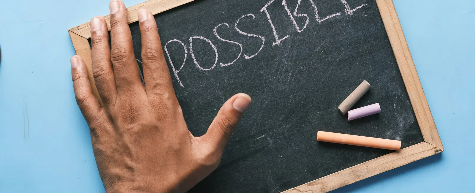 Hand erasing 'im' from 'impossible' on chalkboard to reveal the word 'possible' with chalk pieces nearby.