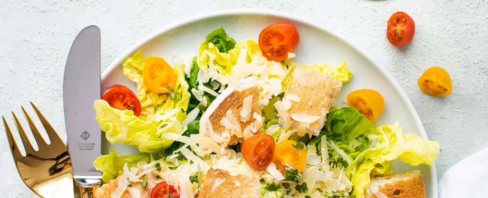 Fresh salad with lettuce, cherry tomatoes, grated cheese, and bread croutons on a white plate with gold cutlery.