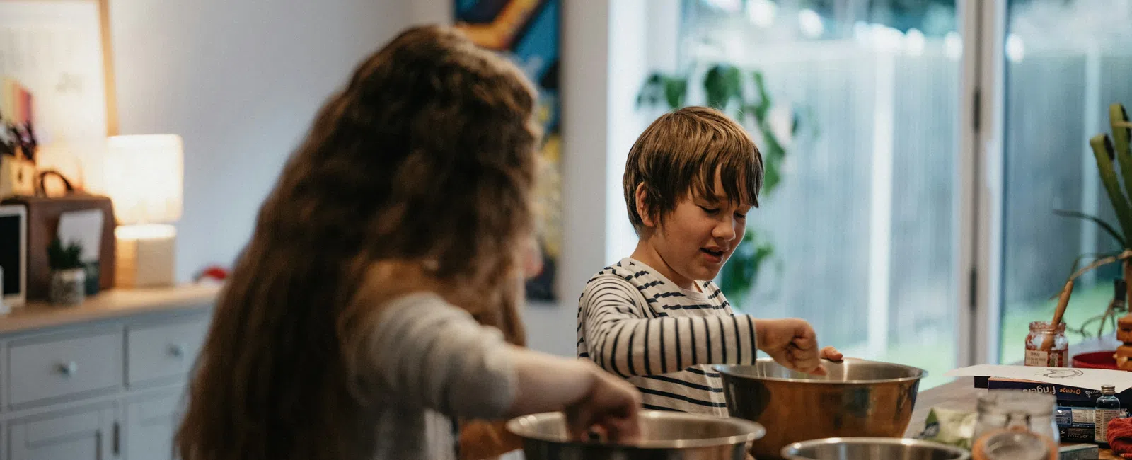 Two children mixing ingredients in metal bowls while baking in a cozy kitchen with natural light.