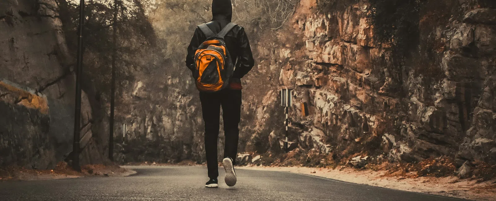 Person with orange backpack walking on a road surrounded by rocky cliffs and trees during dusk.