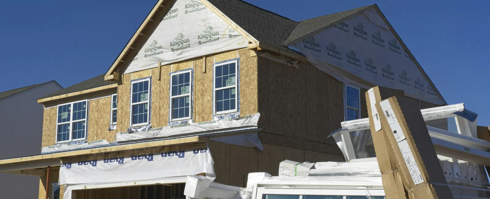 Two-story house under construction with window frames and building materials stacked outside on a clear sunny day.