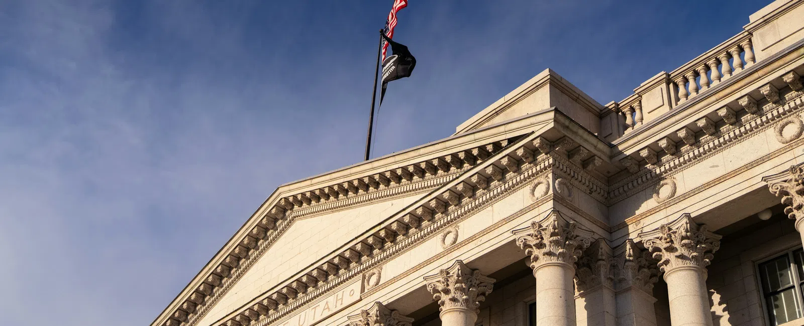 Classical Utah State Capitol building with columns and flags under a partly cloudy blue sky.