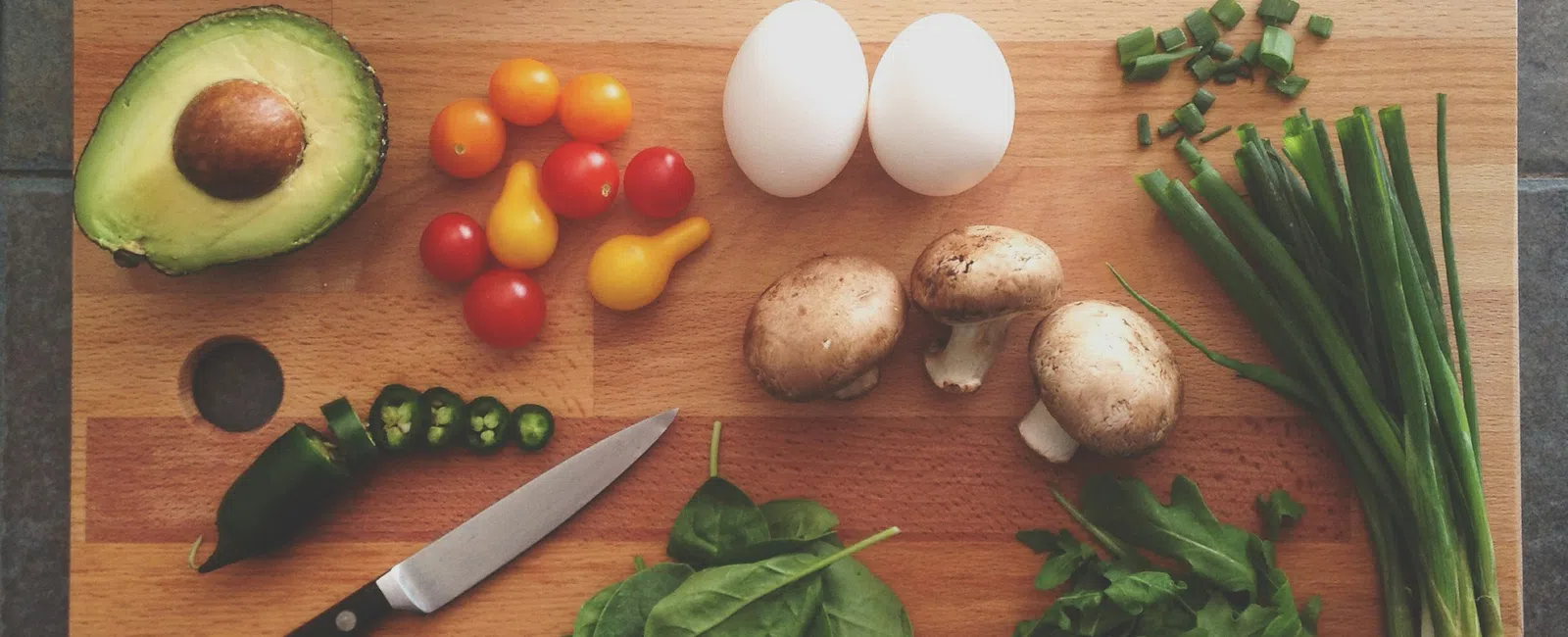 Chopping board with avocado, cherry tomatoes, eggs, mushrooms, green onions, spinach, herbs, and knife above tiled surface.