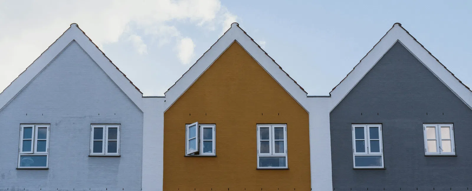 Three adjacent modern houses with gabled roofs in white, mustard yellow, and gray under a partly cloudy sky.
