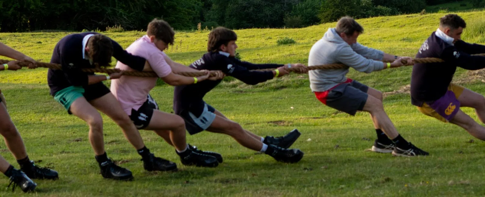 Group of young men playing tug of war on a grassy hill with scenic countryside in the background.
