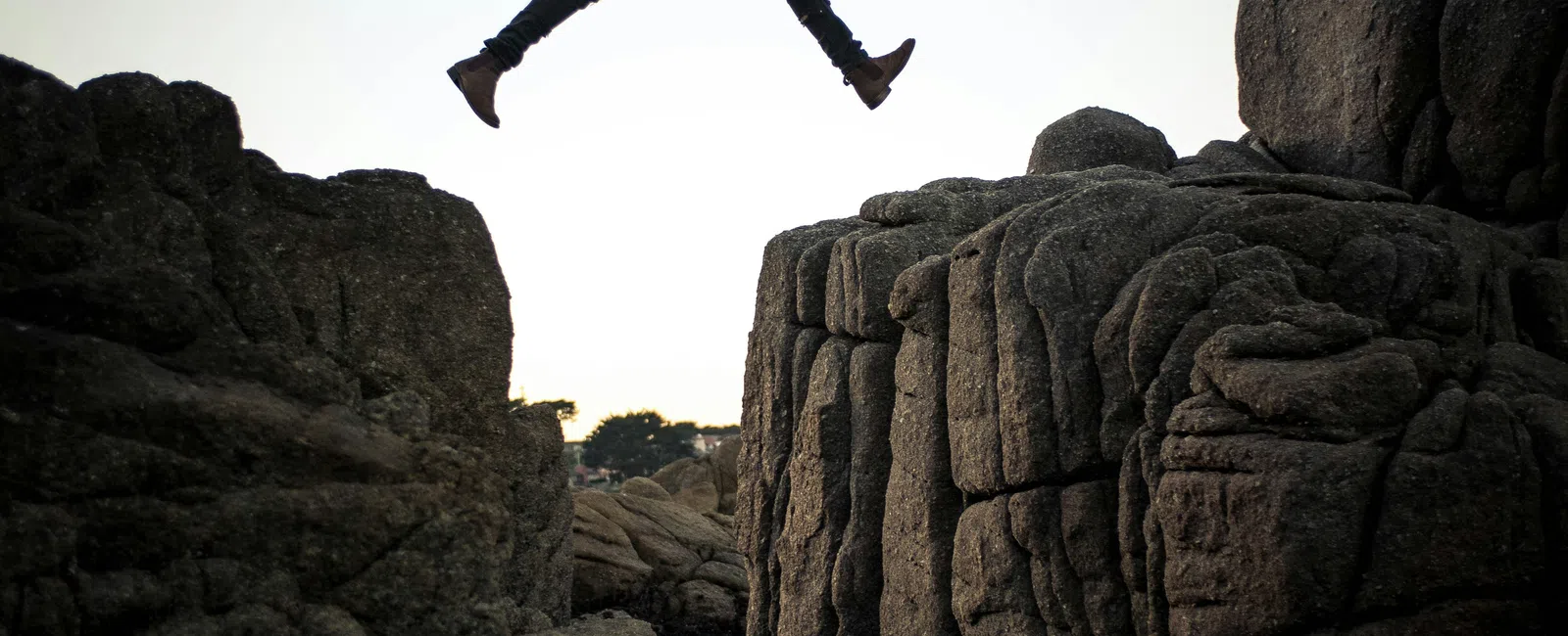 Person jumping across a deep rocky canyon at sunset wearing a brown jacket and ripped jeans.
