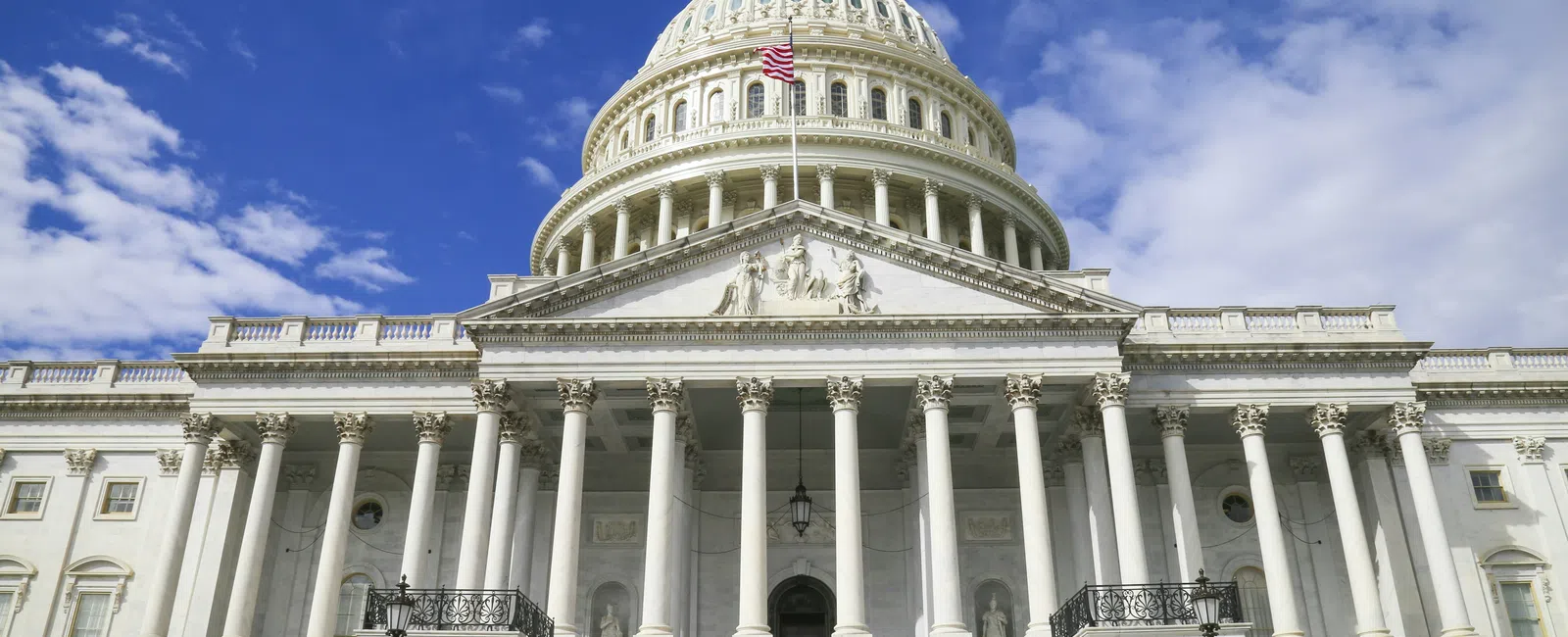 Front view of the United States Capitol building with its dome, columns, and American flag under a blue sky.