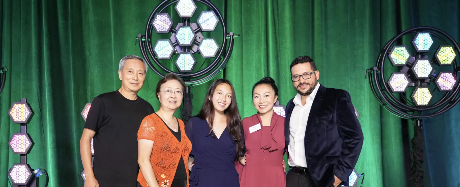 Five people standing together on stage with green backdrop and hexagonal LED lights, smiling at the camera