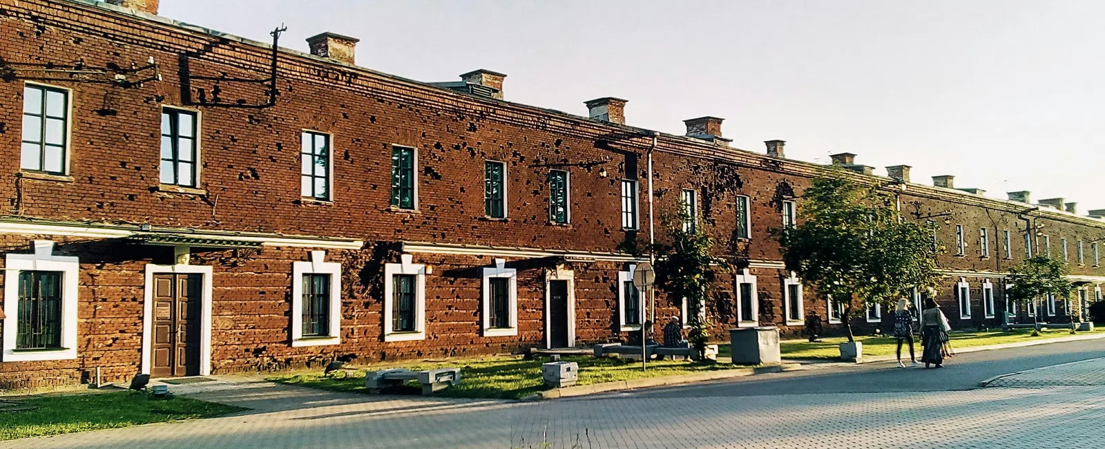 Long brick building with many windows and doors along a paved street, with green lawns and trees in the foreground.