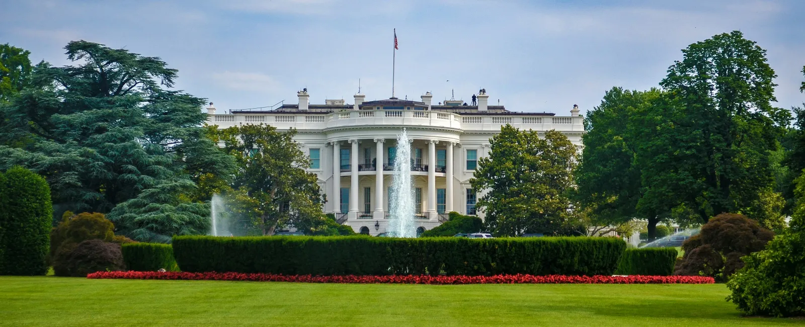 The White House with fountains, green lawns, and trees under a blue sky with clouds